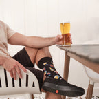 Man in a cream knit polo sits in a slatted patio chair holding a pint of beer toward the table, wearing Get Your Beer Glasses socks in Black with black slip-on sneakers. [lf]