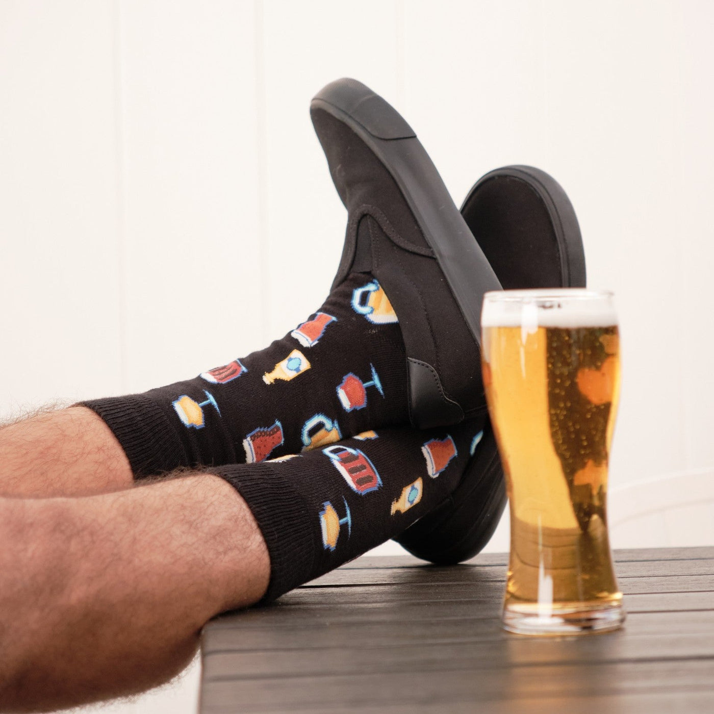 Vertical shot of relaxed feet resting on a wooden table next to a foamy pint glass, featuring Get Your Beer Glasses socks in Black and black slip-on sneakers. [lf]