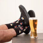 Vertical shot of relaxed feet resting on a wooden table next to a foamy pint glass, featuring Get Your Beer Glasses socks in Black and black slip-on sneakers. [lf]