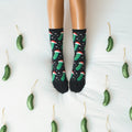 Symmetrical overhead frame of feet pointed up in Christmas Pickle socks in Black, flanked by pickle ornaments on either side of the white bedding. [lf]