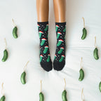 Symmetrical overhead frame of feet pointed up in Christmas Pickle socks in Black, flanked by pickle ornaments on either side of the white bedding. [lf]