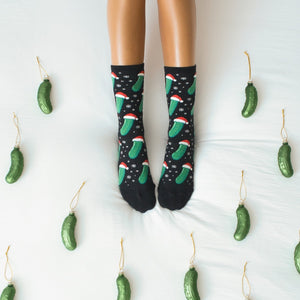 Symmetrical overhead frame of feet pointed up in Christmas Pickle socks in Black, flanked by pickle ornaments on either side of the white bedding. [lf]
