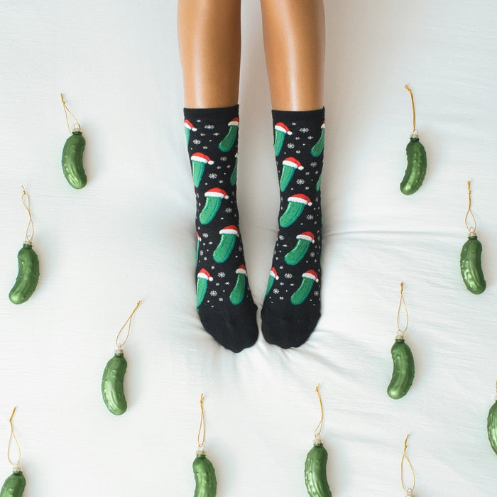 Symmetrical overhead frame of feet pointed up in Christmas Pickle socks in Black, flanked by pickle ornaments on either side of the white bedding. [lf]