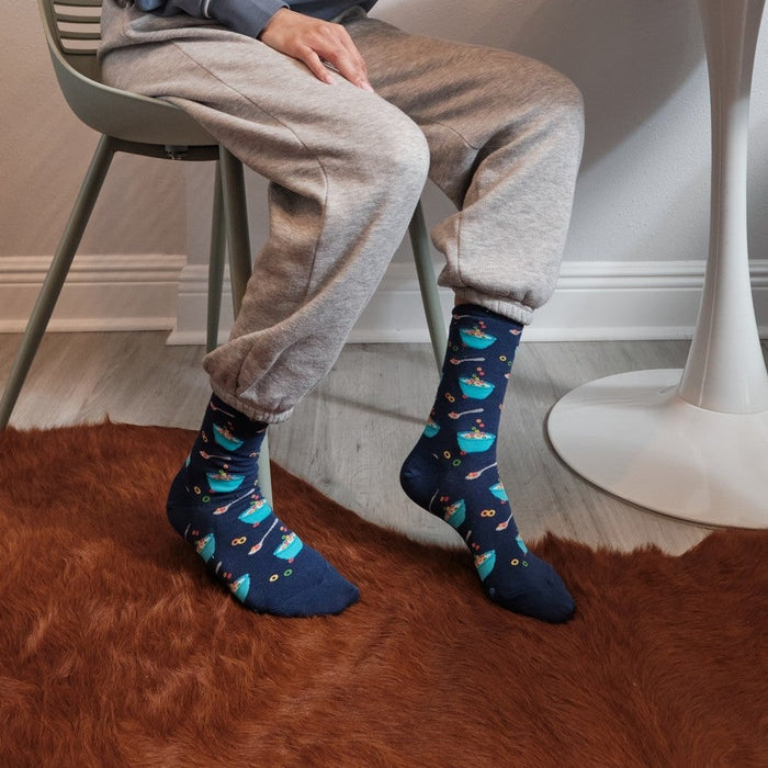 Woman eats cereal at a tulip table on a brown cowhide rug under a giraffe print, wearing Fruity Cereal socks in Navy. [lf]