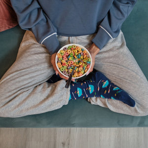 Overhead view of crossed legs on a green velvet couch holding a bowl of fruit loops, feet in Fruity Cereal socks in Navy. [lf]