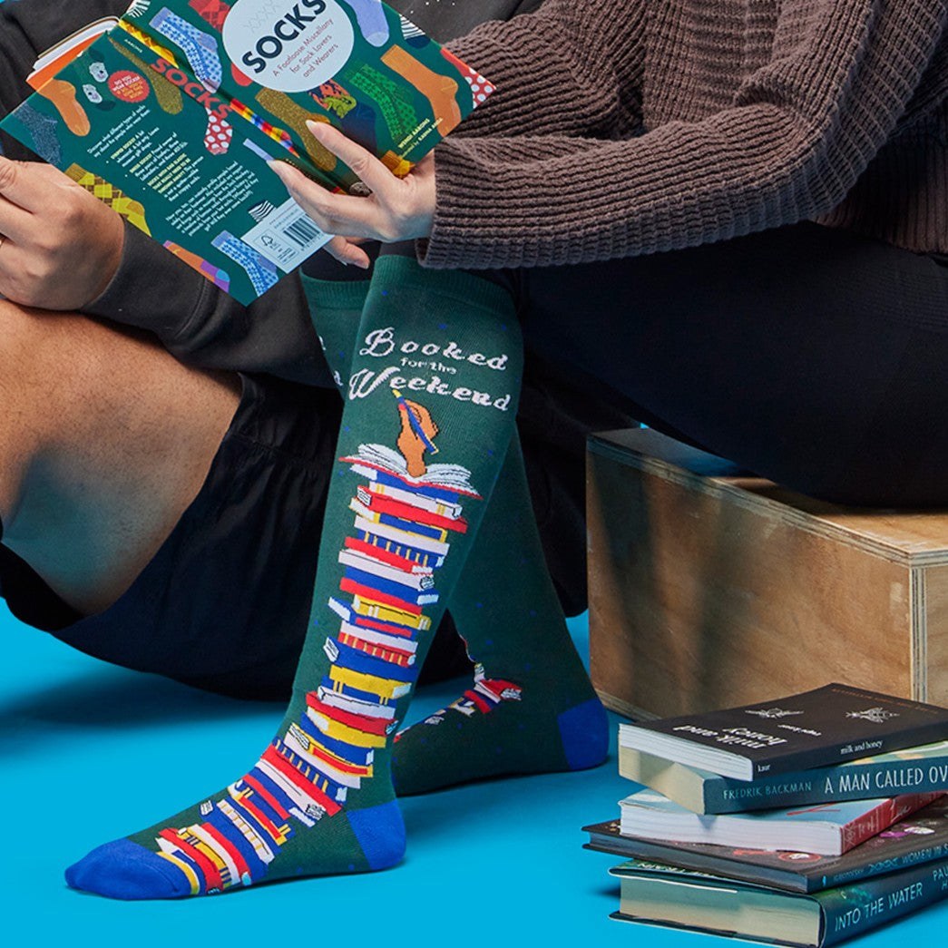 [lf] People relaxing, reading books, wearing green "Booked for the Weekend" socks with colorful book stacks.