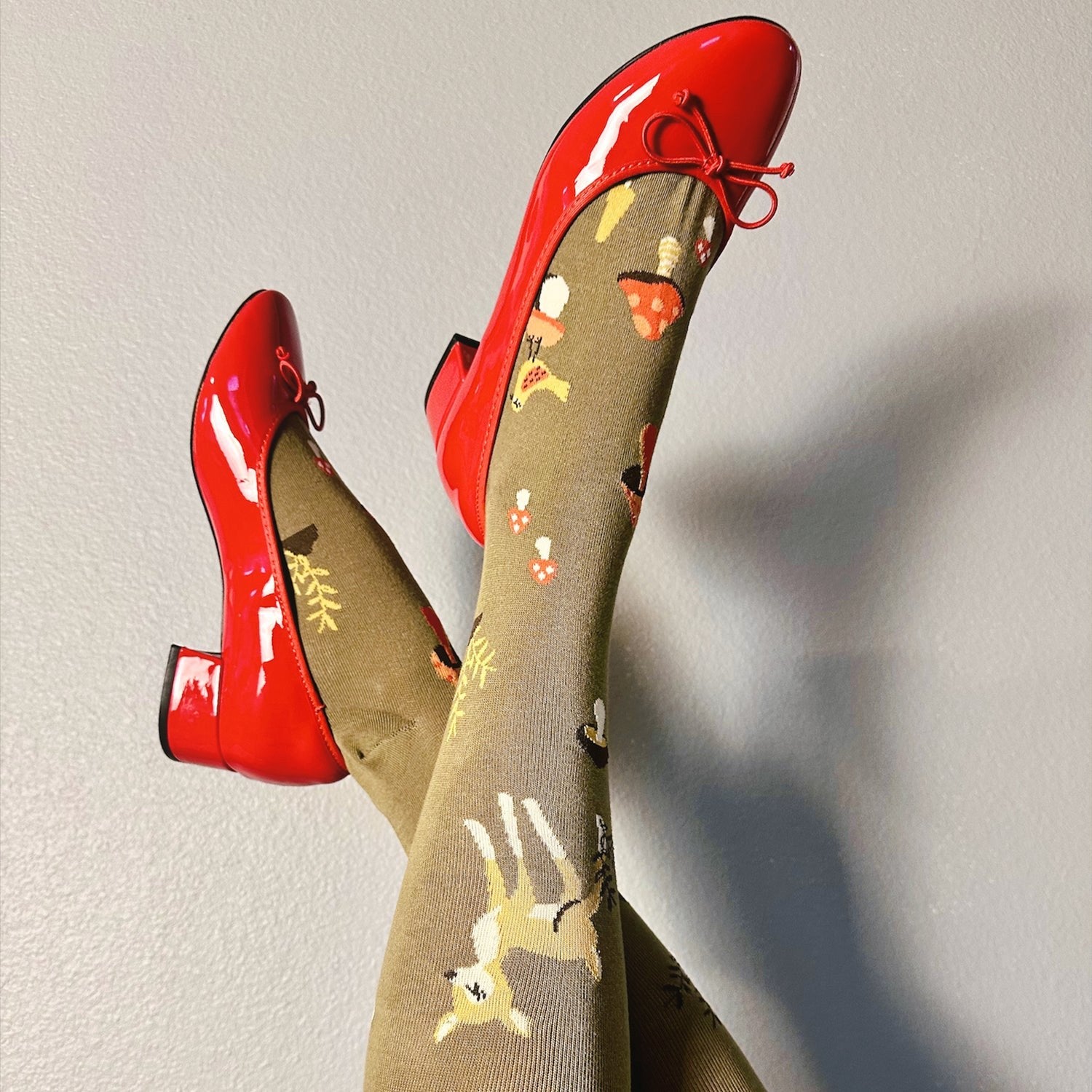 [lf] Legs crossed, a model wears red patent shoes and olive socks with deer and mushroom patterns.