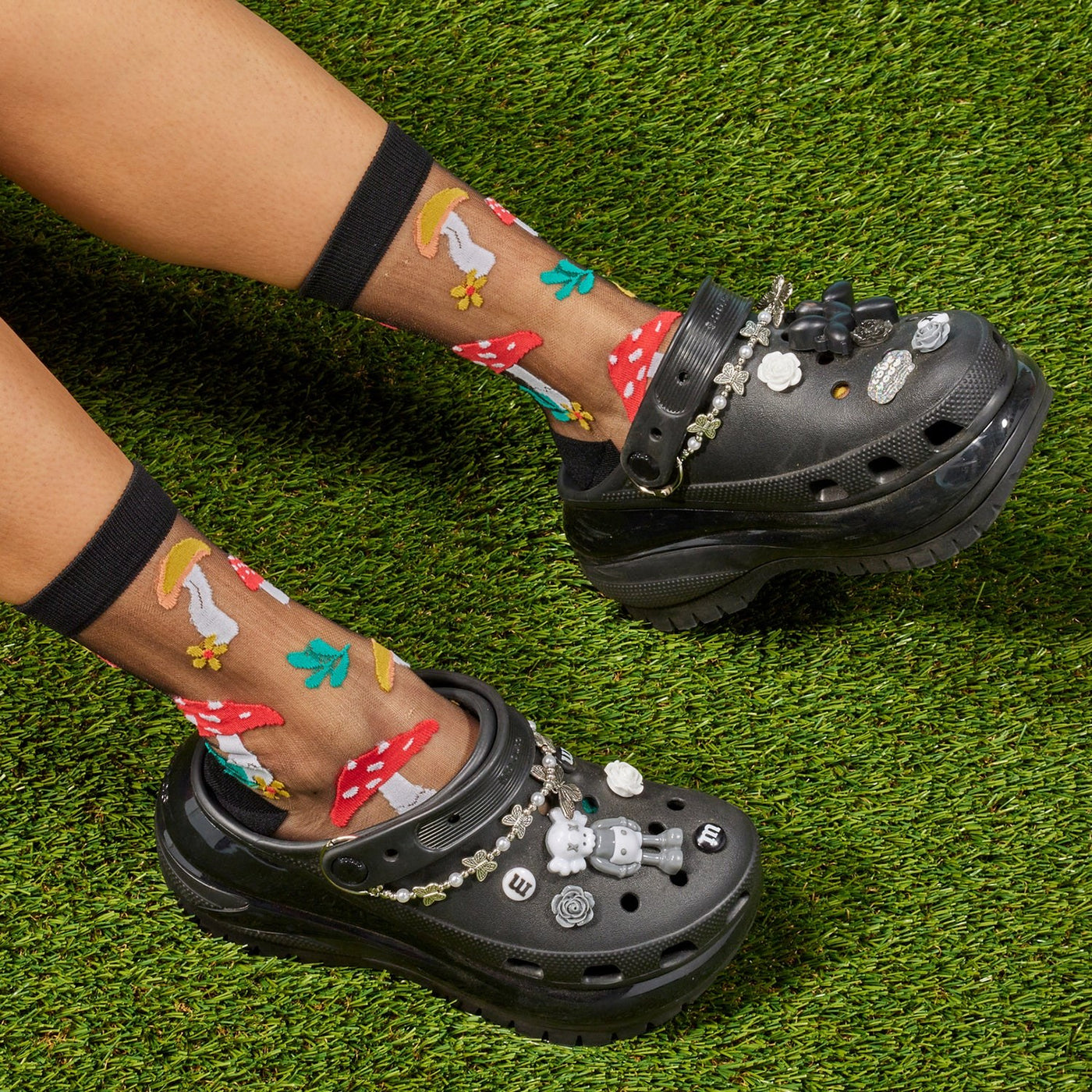 [lf] Legs crossed on grass, model wears sheer black socks with colorful mushroom patterns.
