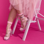 [lf] Model seated, showcasing sheer pink floral socks with clear heels against a vibrant pink background.