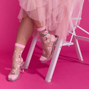 [lf] Model seated, showcasing sheer pink floral socks with clear heels against a vibrant pink background.