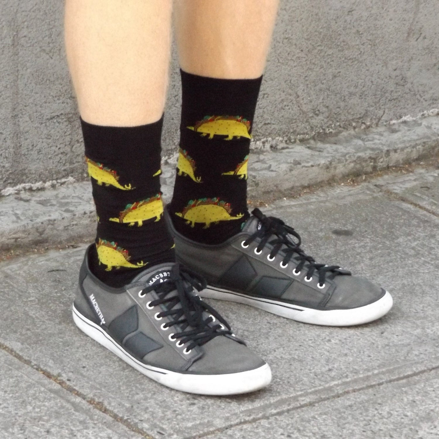 [lf] Person standing on pavement wearing black socks with yellow taco-saurus pattern.