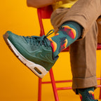 [lf] Person in green sneakers and brown pants, showing colorful mountain-patterned socks against a yellow background.