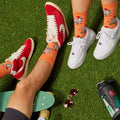 [lf] Legs on artificial grass, wearing orange socks with skateboarding chickens and red and white sneakers.
