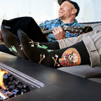 [lf] Man laughing, playing guitar, feet up by fire pit, wearing black lumberjack socks.