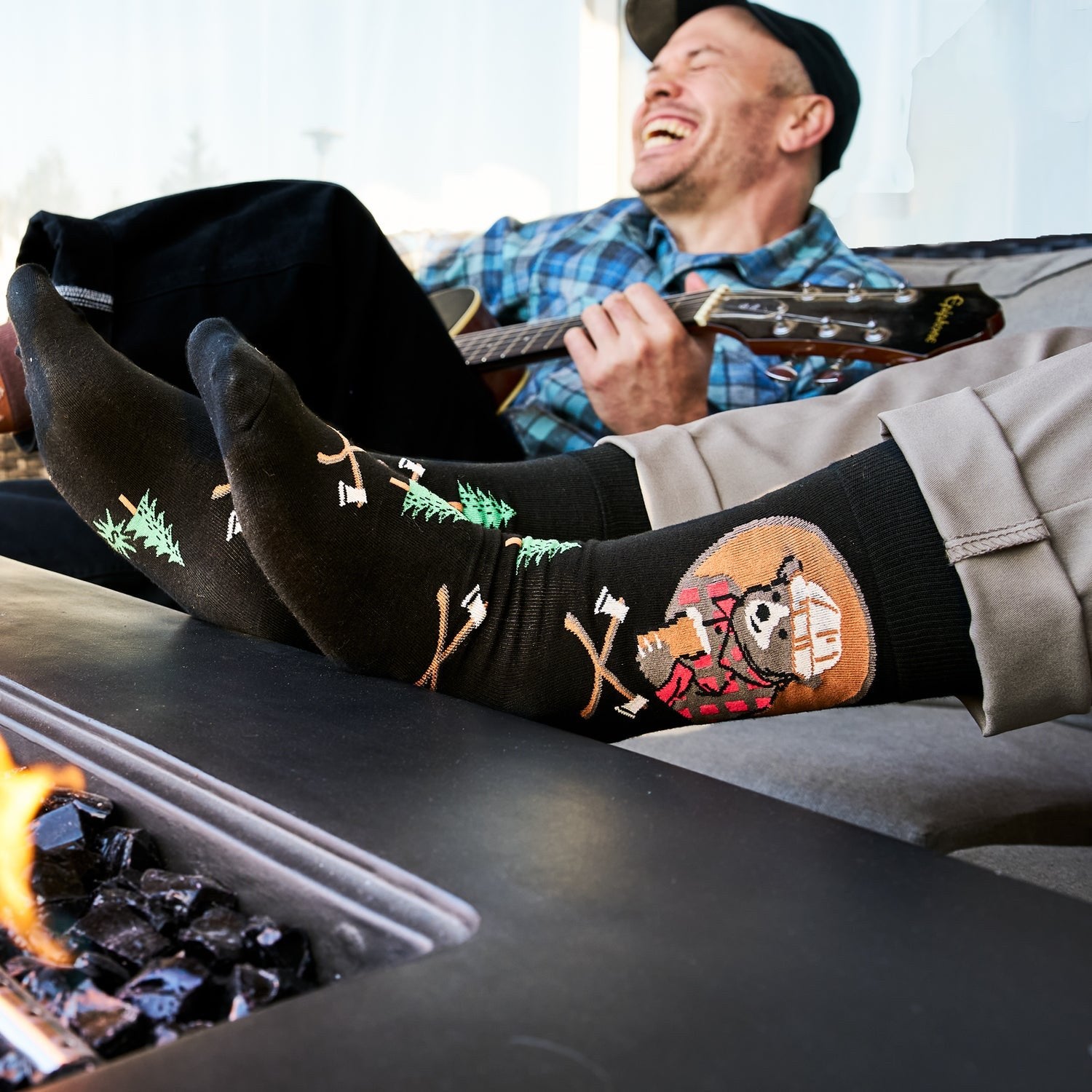 [lf] Man laughing, playing guitar, feet up by fire pit, wearing black lumberjack socks.