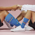 [lf] Two people sit holding hands, wearing blue socks with rainbow "QUEER JOY" text and yellow stars.