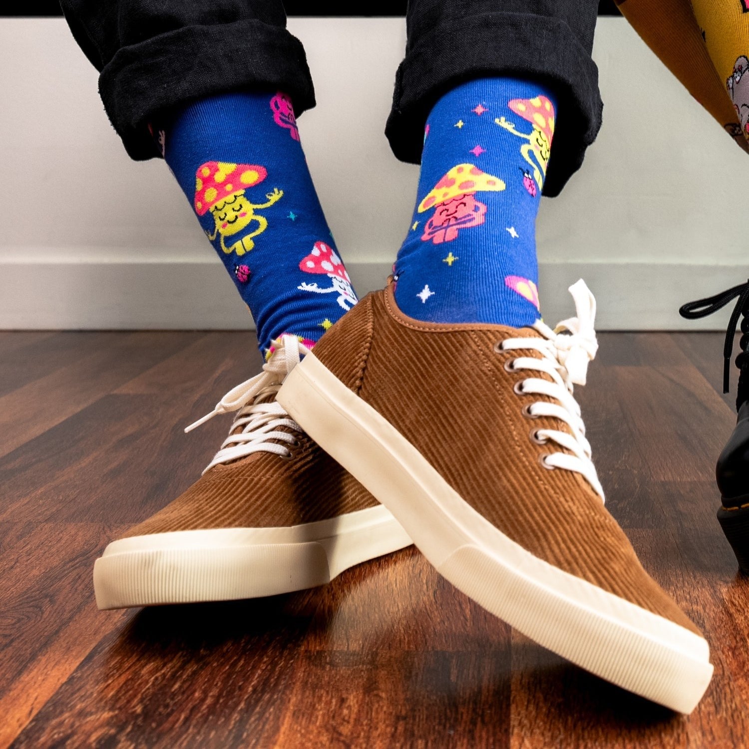 [lf] Person wearing black pants, brown corduroy shoes, and blue mushroom-patterned socks on a wooden floor.