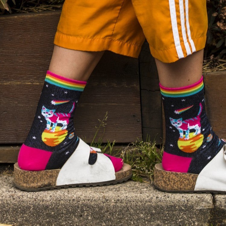 [lf] Person in orange shorts and sandals wearing black socks with rainbow cuffs and space cat design.