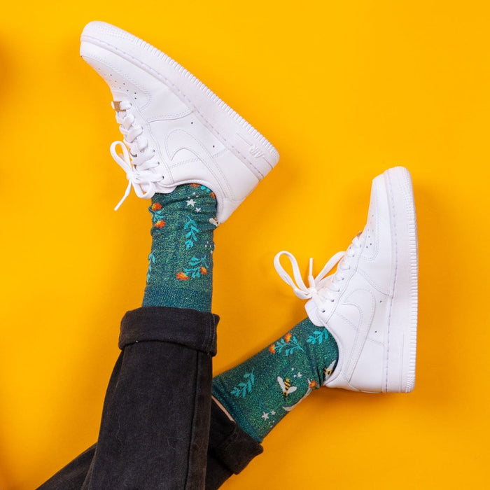[lf] Legs in black jeans and white sneakers, showcasing green socks with floral and bee patterns.