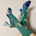 [lf] Legs crossed, wearing green beetle-patterned socks and metallic heels against a light background.