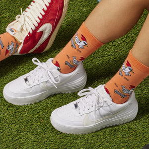 [lf] Legs in orange chicken-on-skateboard socks and white sneakers on green turf.