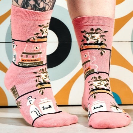 [lf] Pink socks with cats, plants, and books, worn by a person standing on a patterned floor.