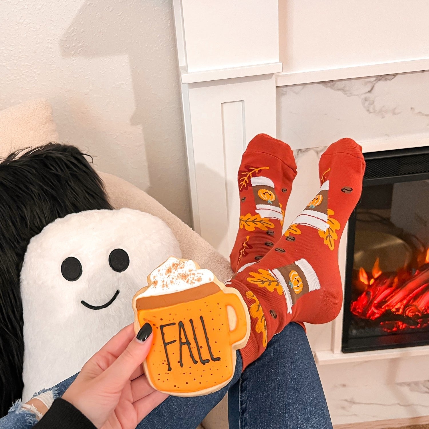 [lf] Cozy scene with feet in red fall-themed socks, holding a "FALL" cookie by a fireplace.