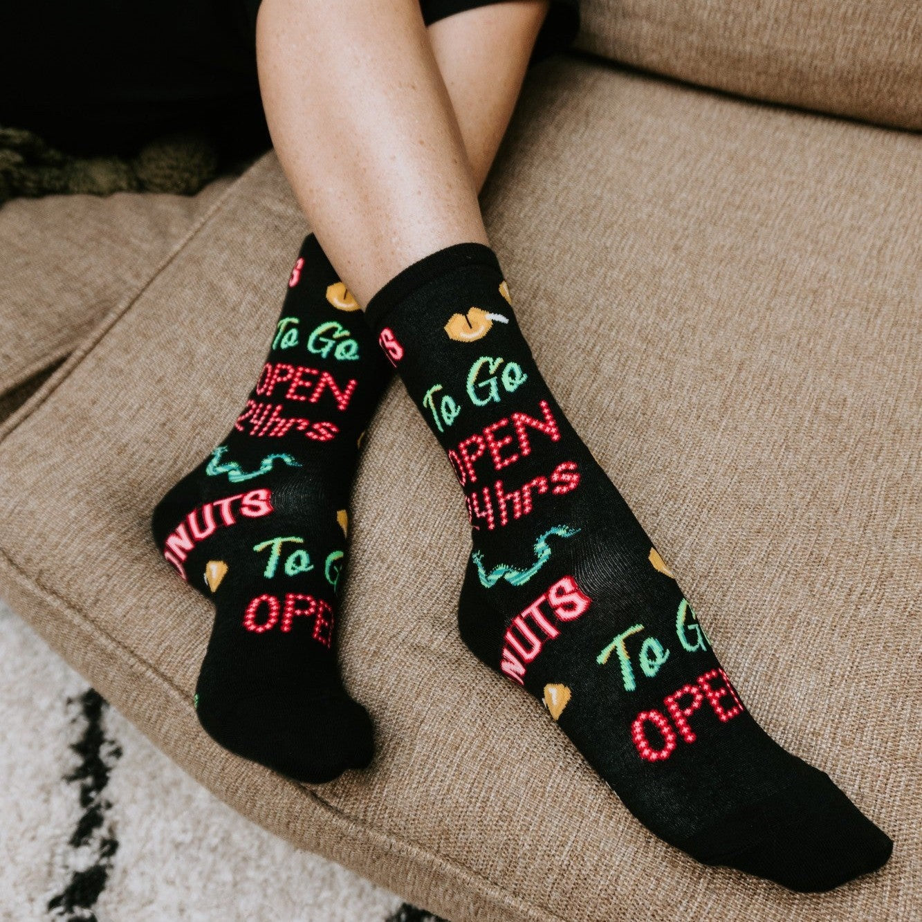 Close detail of crossed ankles on a beige cushion highlighting the To Go, Open 24 Hrs and donut prints on the 24 Hour Diner socks. [lf]