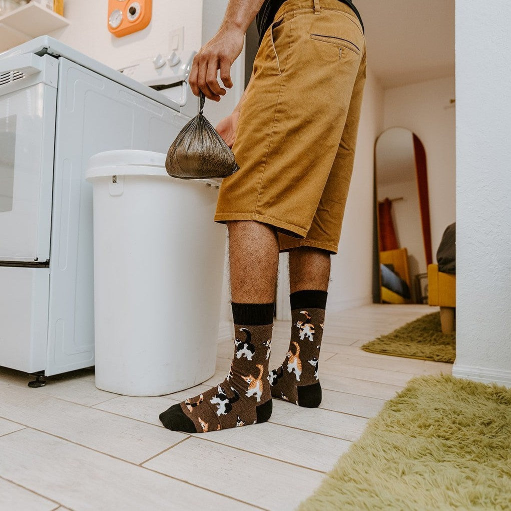 Holding a small trash bag inside an apartment, the wearer steps past a green shag rug while their Cat Butts socks in Brown Heather pop against pale tile, kitty tails arched mid-strut. [lf]