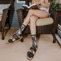 A woman reads on a wicker porch chair in Corgi Butt grey socks with adorable corgi prints paired with brown leather Birkenstocks. [lf]