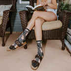 A woman reads on a wicker porch chair in Corgi Butt grey socks with adorable corgi prints paired with brown leather Birkenstocks. [lf]