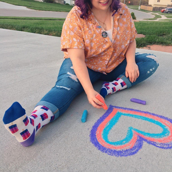 A woman sits on a driveway drawing a rainbow heart in chalk, wearing Bisexual Pops socks with pink, purple, and red popsicle prints on a blue knit. [lf]