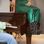 A red-shirted woman in a shiny green skirt sits atop the grand piano holding a glass of wine, her black Christmas Cocktails socks lined with festive cocktail glasses as her partner plays in the foreground. [lf]
