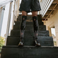Spider Webs socks shown on a model standing on a sunlit garden staircase. [lf]
