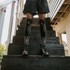 Spider Webs socks shown on a model standing on a sunlit garden staircase. [lf]