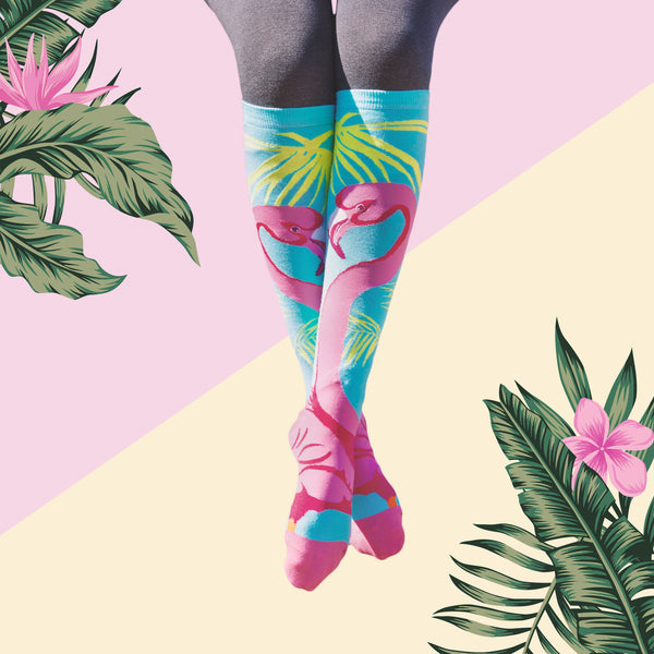 Lifestyle shot of model's legs wearing the flamingo knee-high socks against a tropical-themed background. [lf]