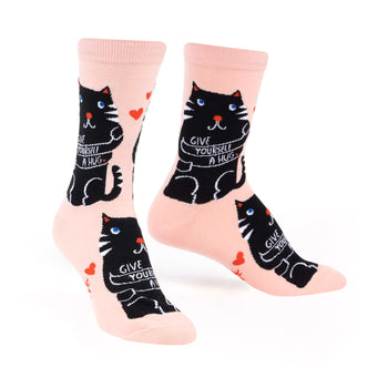 Crew socks in pale pink with a repeating pattern of black cats hugging themselves and the words 'Give Yourself A Hug'.