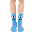 Fuzzy slipper socks in light blue with a playful monster face design and purple accents. Non-skid grips are visible on the soles. [lf]