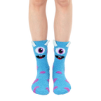 Fuzzy slipper socks in light blue with a playful monster face design and purple accents. Non-skid grips are visible on the soles. [lf]