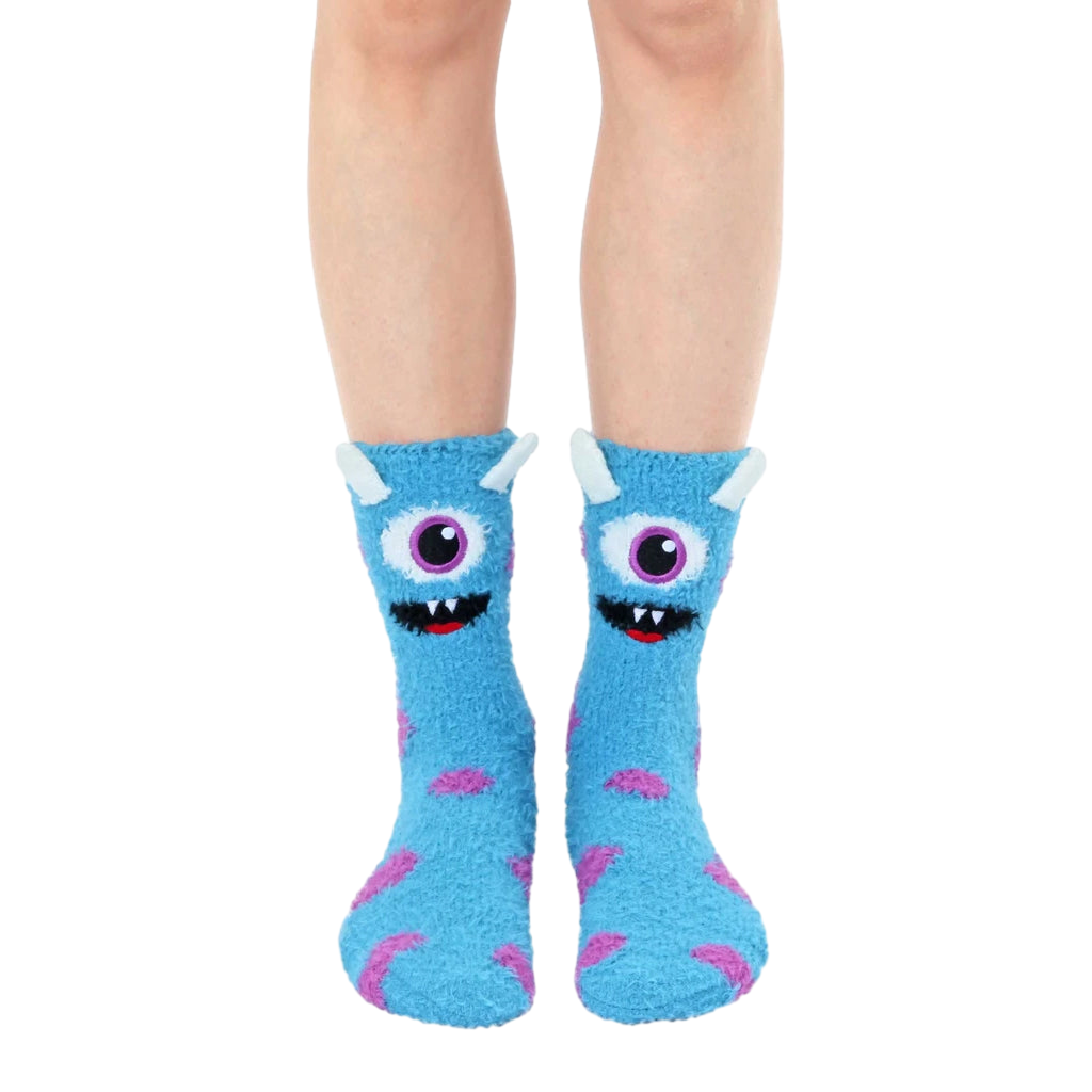 Fuzzy slipper socks in light blue with a playful monster face design and purple accents. Non-skid grips are visible on the soles. [lf]