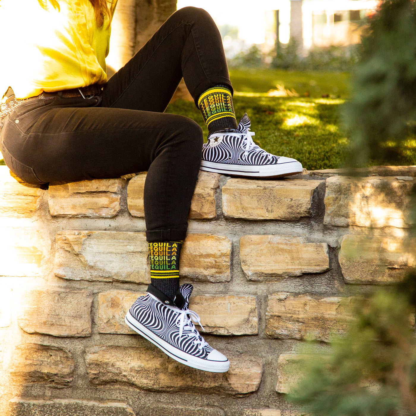 Lifestyle shot of a person wearing black jeans and a yellow shirt, showing off the Tequila socks with black and white zebra print shoes. [lf]