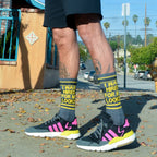 Lifestyle shot showing a person wearing the 'I Was Hired For My Looks' socks with Adidas sneakers. The socks are dark gray with yellow and black stripes and text. [lf]