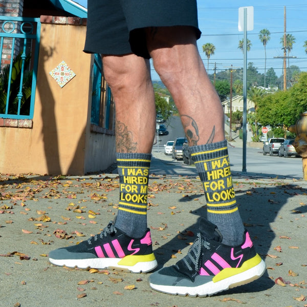 Lifestyle shot showing a person wearing the 'I Was Hired For My Looks' socks with Adidas sneakers. The socks are dark gray with yellow and black stripes and text. [lf]