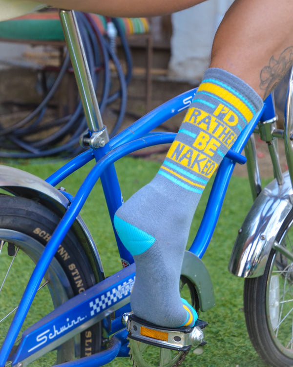 Lifestyle shot of a person's leg wearing a 'I'd Rather Be Naked' sock while sitting on a blue bicycle. [lf]