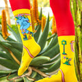 Lifestyle shot: Model wearing yellow Skate Day socks with red pants against a green background. [lf]