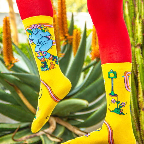 Lifestyle shot: Model wearing yellow Skate Day socks with red pants against a green background. [lf]