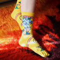 Lifestyle shot: Close-up of feet in yellow Skate Day socks on an orange and brown rug. [lf]