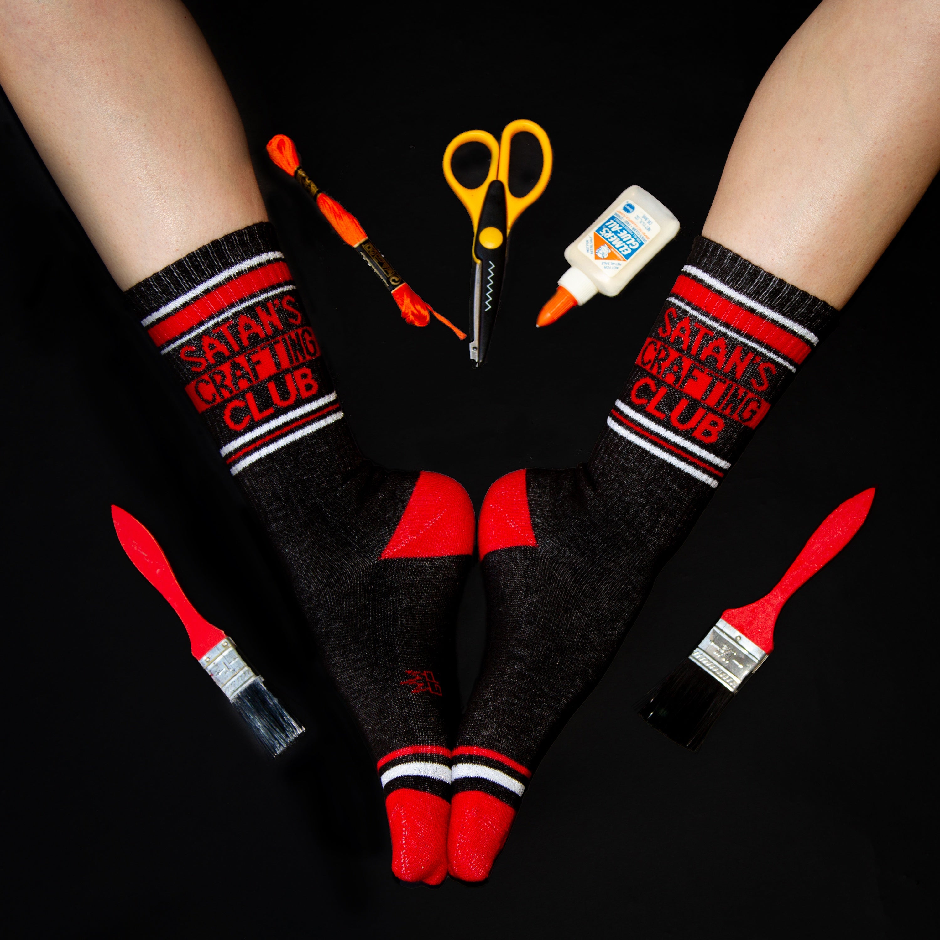 Lifestyle shot of a pair of Satan's Crafting Club socks worn with crafting supplies arranged around them. [lf]