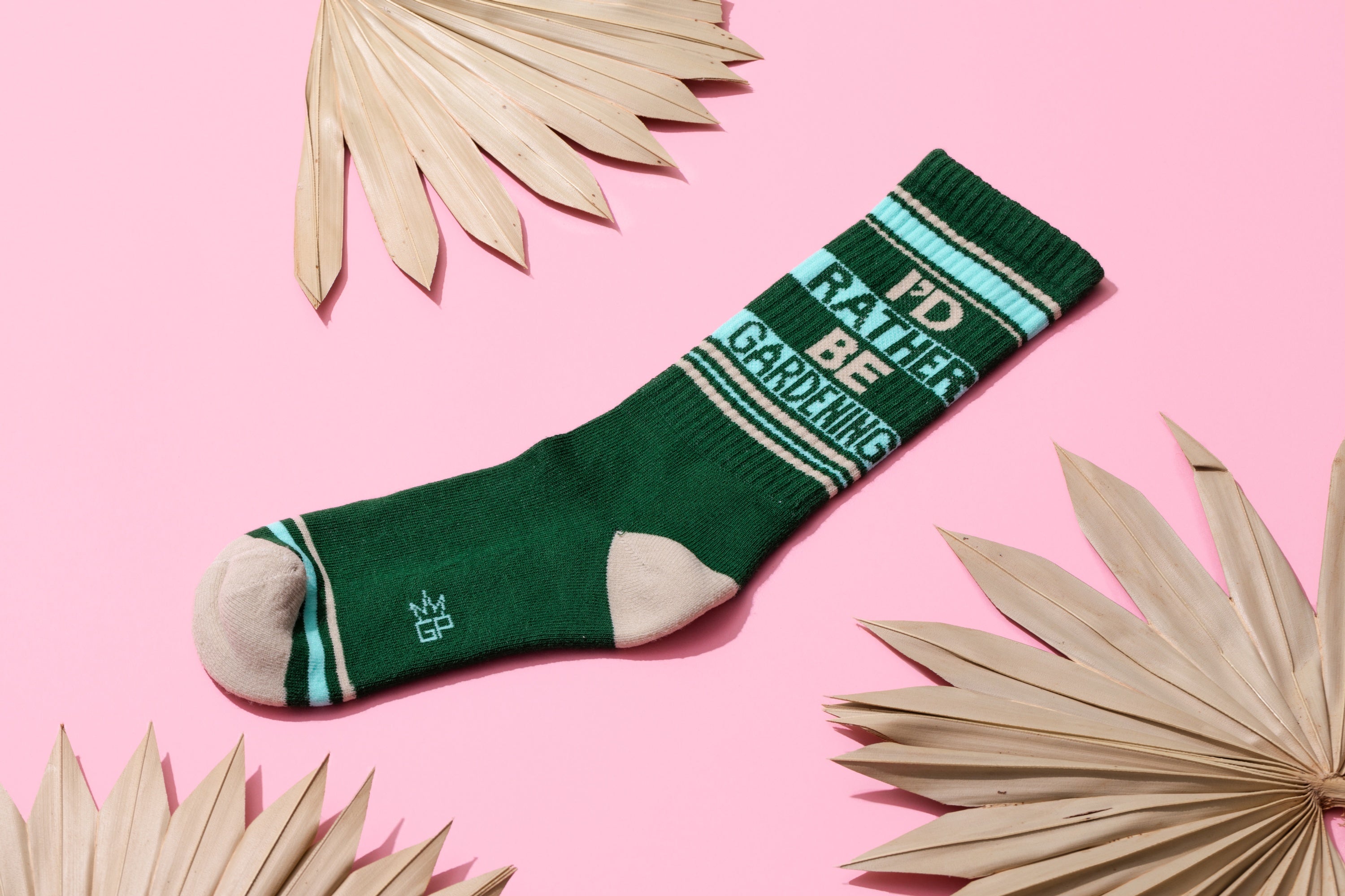 Flat lay product shot of a single 'I'd Rather Be Gardening' sock on a pink background with palm leaves. [lf]