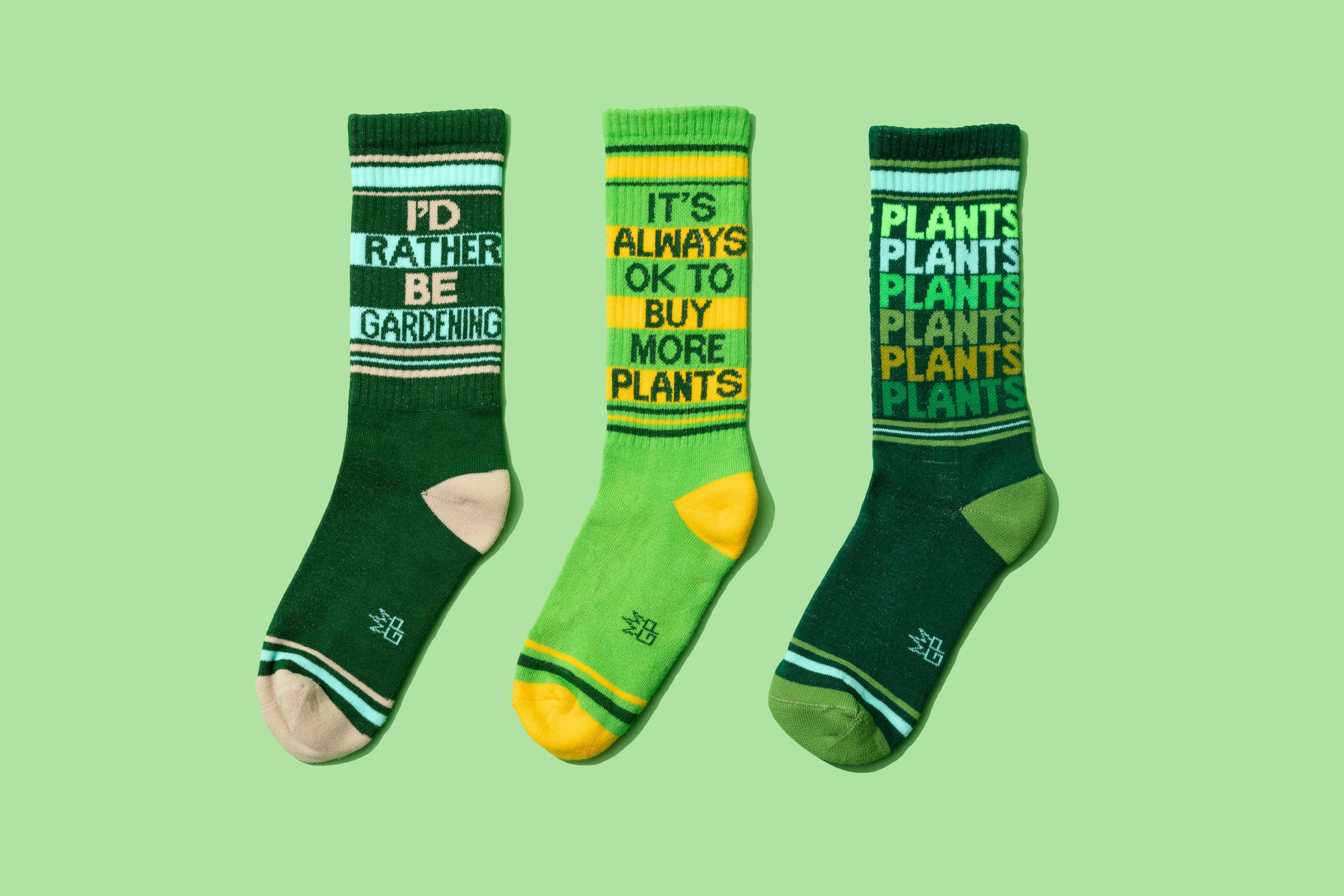 Product shot of three different gardening-themed socks: 'I'd Rather Be Gardening', 'It's Always Ok To Buy More Plants', and 'Plants Plants Plants'. [lf]